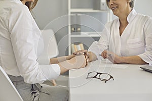 Senior doctor holding patient`s hand, showing support at clinic, closeup. Young woman on visit to physician at hospital