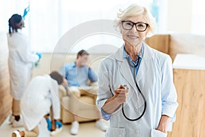 Senior doctor in lab coat and glasses smiling cheerfully and holding stethoscope