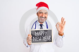 Senior doctor man wearing christmas hat holding blackboard over isolated white background doing ok sign with fingers, excellent