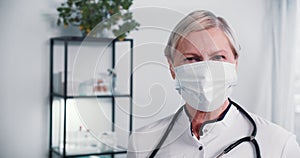 Senior doctor posing at work. Happy 60 grey haired physician woman wears face mask, white lab coat and stethoscope.