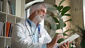 Senior Doctor Thinking About Patient's Diagnosis and Typing on a Tablet Computer. His Office is Bright and Modern.