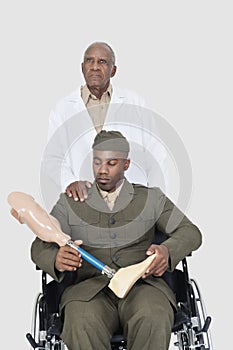 Senior doctor with US military officer holding artificial limb as he sits in wheelchair over gray background