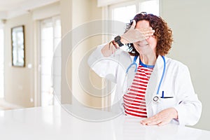 Senior doctor woman wearing medical robe at the clinic smiling and laughing with hand on face covering eyes for surprise
