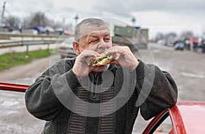 Senior driver gobbling lyulya kebab in lavash near his car