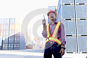 A senior elderly Asian worker engineer wearing safety vest and helmet standing and holding radio walkies talkie at logistic