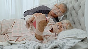Senior elderly couple wearing pyjamas lying on bed looking on mobile phone laughing and having fun