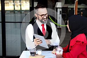 Senior elderly mature business man using tablet and discussing with hijab Muslim women during having a coffee at outdoor cafe,