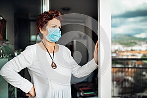 Senior elderly sad woman at home,wearing mask on balcony window.Coronavirus COVID-19 disease outbreak infection risk.Lockdown