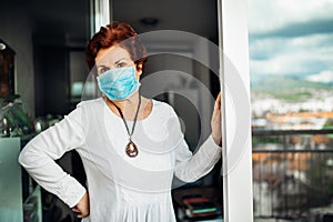Senior elderly sad woman at home,wearing mask on balcony window.Coronavirus COVID-19 disease outbreak infection risk.Lockdown