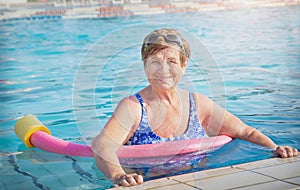 Senior (elderly) woman (over age of 50) in sport goggles, swimsuit and with swim noodles in swimming pool.