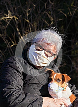 Senior elderly woman wearing mask sitting in park with her dog wearing simple respirator as well. Pets are not vulnerable to