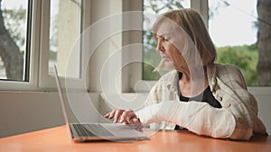 Senior elderly woman working at home with laptop computer with injury broken arm in cast indoors in front of windows
