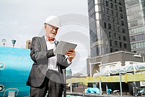 Senior engineer man using tablet to check and maintenance in area of construction site with other building as background