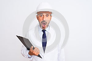 Senior engineer man wearing security helmet holding clipboard over isolated white background scared in shock with a surprise face,