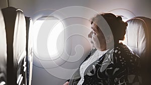 Senior European female airplane passenger sitting on the airplane window seat, nervous and scared to fly, looking around