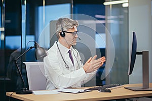 Senior and experienced doctor on video communication consults the patient, uses a headset, the man works in office of clinic