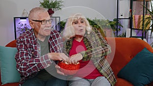 Senior family couple grandparents man woman watching interesting TV football movie, eating popcorn