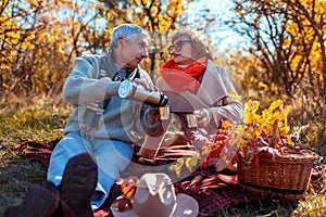 Senior family couple having tea pouring it from thermos in fall forest. Happy man and woman enjoying picnic outdoors