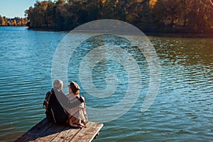 Senior family couple relaxing by autumn lake. Happy man and woman enjoying nature and hugging sitting on pier