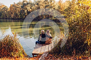 Senior family couple relaxing by autumn lake. Man and woman enjoying nature and hugging sitting on pier