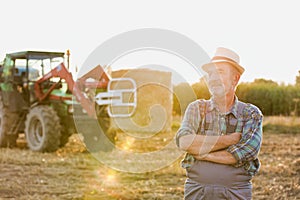 Senior farmer standing with arms corssed against farm tractor, round bale hay in field with yellow lens flare in background photo