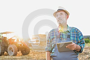 Senior farmer standing with arms corssed against farm tractor, round bale hay in field with yellow lens flare in background photo