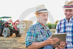 Senior farmer standing while using digital tablet with mature farmer against farm tractor, round bale hay in field with y photo