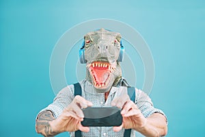 Senior fashion man taking selfie with mobile smartphone while wearing t-rex mask - Hipster guy having fun listening music