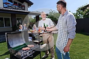 Senior father and adult son drinking beer while grilling meat outdoors