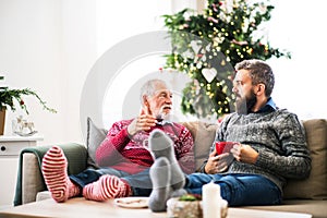 A senior father and adult son sitting on a sofa at home at Christmas time, talking.