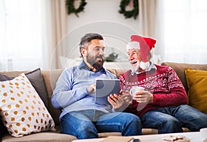 A senior father and adult son with tablet sitting on a sofa at home at Christmas time.
