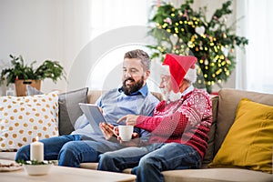 A senior father and adult son with tablet sitting on a sofa at home at Christmas time.