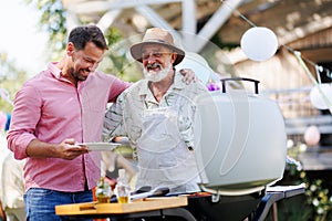 Senior father and mature son grilling together at garden bbq party. Man looking at full plate with grilled food.