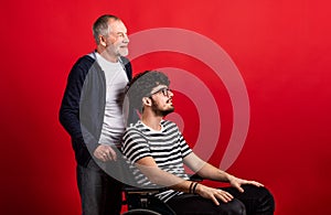 Senior father with young son in wheelchair in a studio on red background.