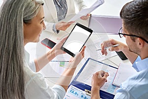 Senior female business leader holding tablet with colleagues in office at desk.