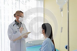 Senior Female doctor wearing protective face mask explaining diagnosis to her Asian patient in medical room
