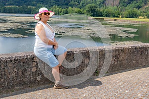 Senior female tourist sitting on fence at shore of Echternach lake, serene expression