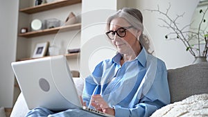 Senior female typing email on laptop, blogging, writing message, working on project from home