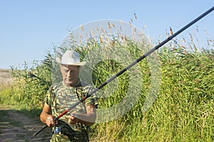 Senior fisherman in hat with a fishing rod prepares to fish on the shore with reeds. An elderly retired man is resting in nature.