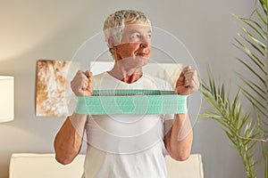 Senior fit grey-haired mature man wearing casual white T-shirt sitting on sofa at home interior having home workout with elastic