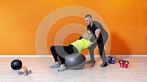 Senior fit woman doing workout exercise siting on a ball at the gym isolated on orange background followed by a pesonal trainer.