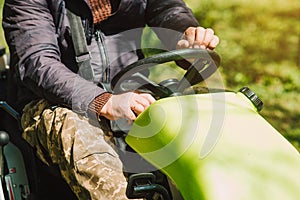 Senior gardener in medical mask driving a riding lawn mower in a garden.