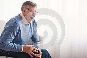 Senior gentleman having knee pain sitting on sofa at home