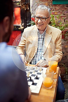 Senior good looking male looking at his younger opponent playing chess, sitting in outdoor cafe