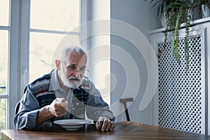 Senior grandfather with grey hair and beard sitting alone in the kitchen eating breakfast