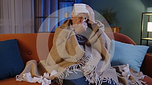 Senior grandfather man suffering from cold or allergy, blows nose snot into napkin sitting at home