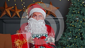 Senior grandfather parodies Santa Claus presenting Christmas gift box, holidays celebration at home