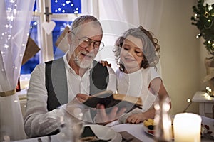 Senior grandfather with small granddaughter indoors at Christmas, reading Bible.