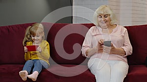 Senior grandmother with child girl granddaughter using digital mobile phone, playing games at home