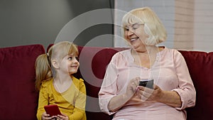 Senior grandmother with child girl granddaughter using digital mobile phone, playing games at home